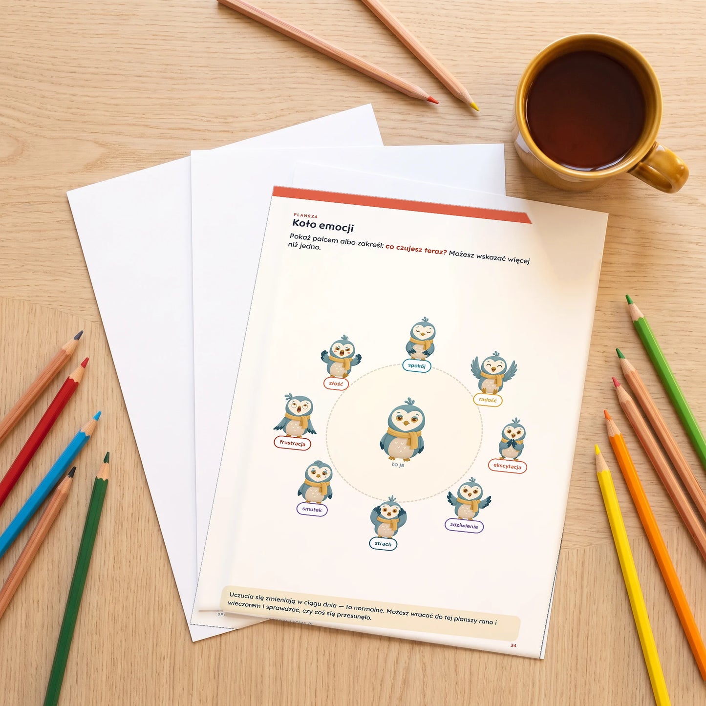 Wydrukowana plansza Koło emocji ze Spokojnej Sowy leżąca na stole obok kredek i kubka herbaty